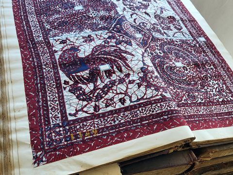 An wax print sample book open on display at Galerie Atiss. Bold colors of indigo and maroon stand out as they surround the image, with a rooster, flowers, and branches in the center of the image broken down into four quadrants.