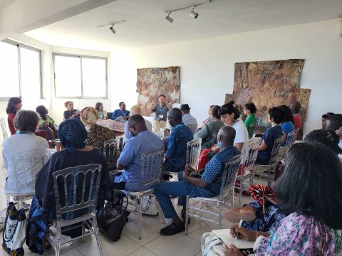 Participants listening to the Archives Beyond The Institutional roundtable at Galerie Atiss, Dakar, Senegal. Everyone is facing the speakers who are laid out in a semi-circle, with a wax print textile book in between both parties.